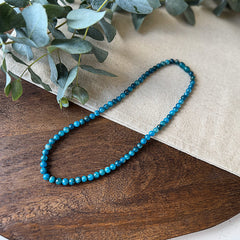 Aventari Apatite Intellect Necklace featuring blue-green beads laid flat on a natural wood and linen surface with eucalyptus accents.